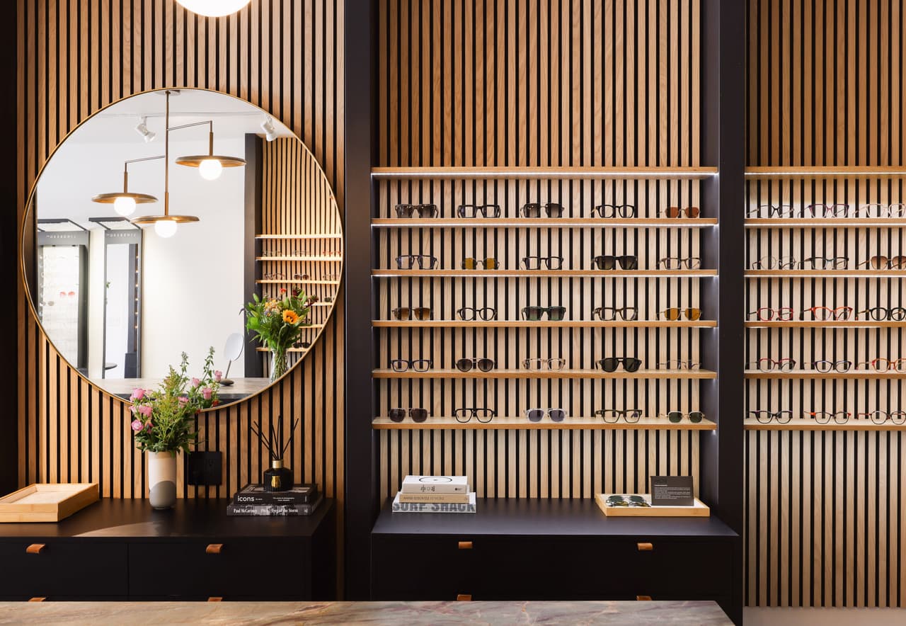 A wall of frames on lit oak shelves beside a large round brass mirror, with flowers and art books on the counter below.